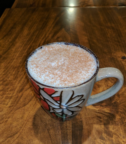 A decorative mug filled with frothy coffee topped with a sprinkle of cinnamon, resting on a wooden table.